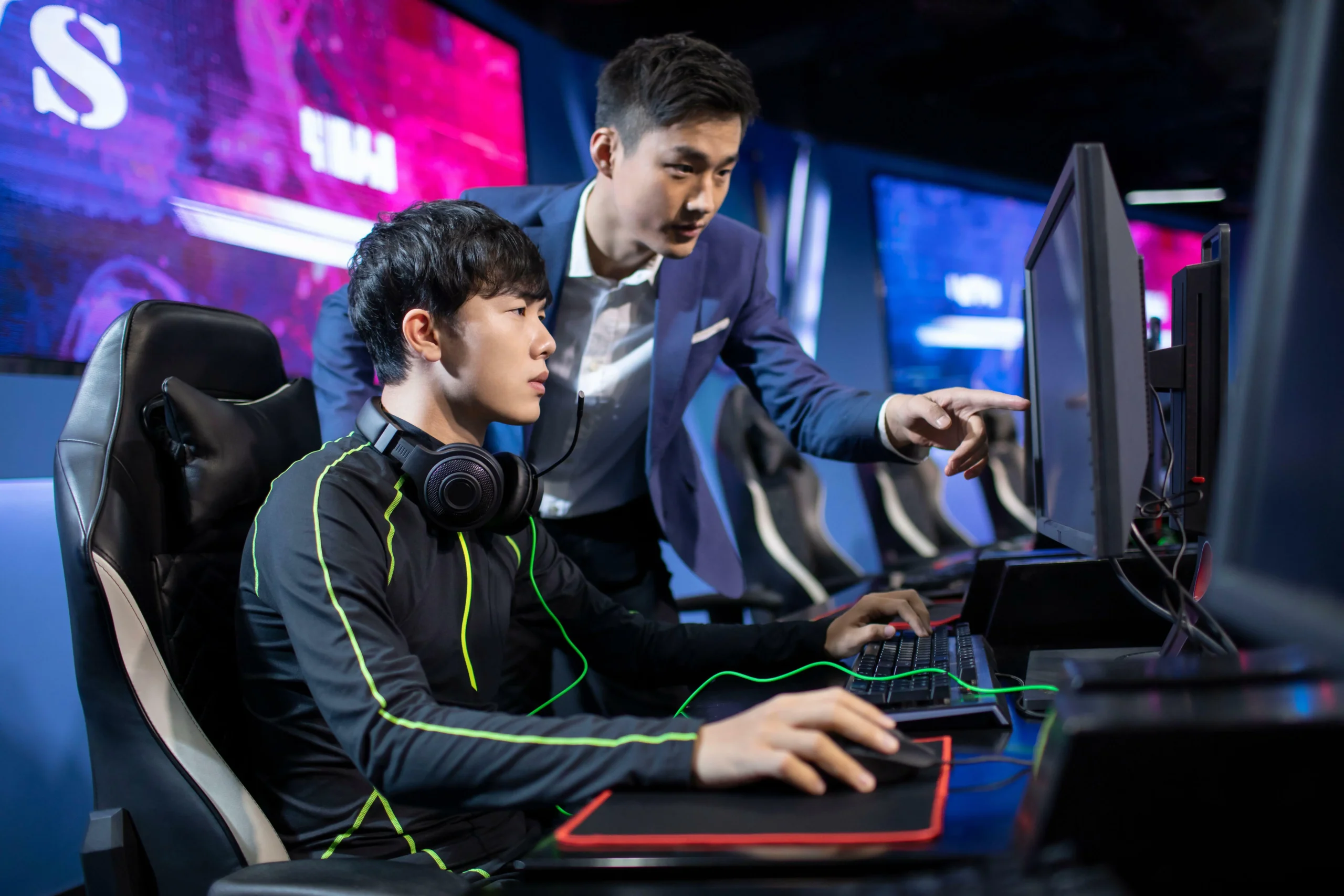 A player in a gaming headset focuses on the screen while a coach in a suit