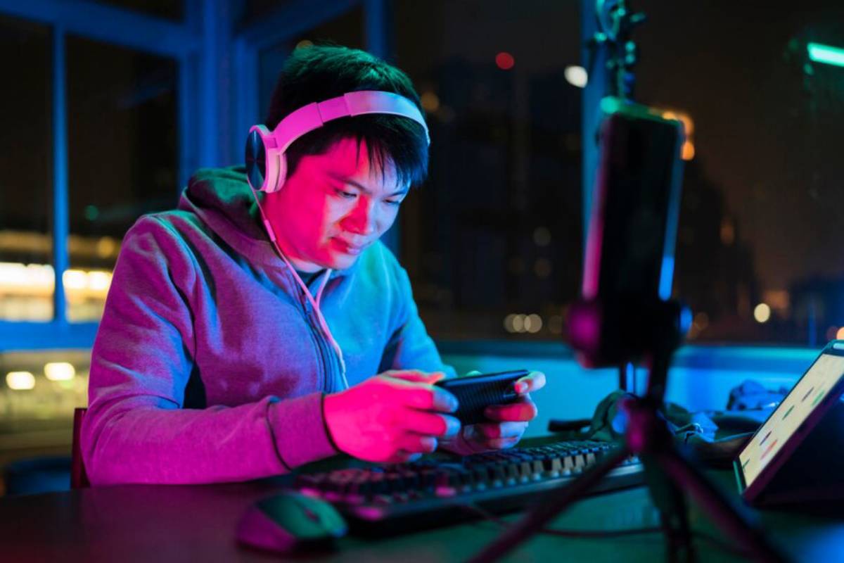 A person in a hoodie sits at a desk with a keyboard and tablet, playing game on mobile, surrounded by colorful ambient lighting at night.