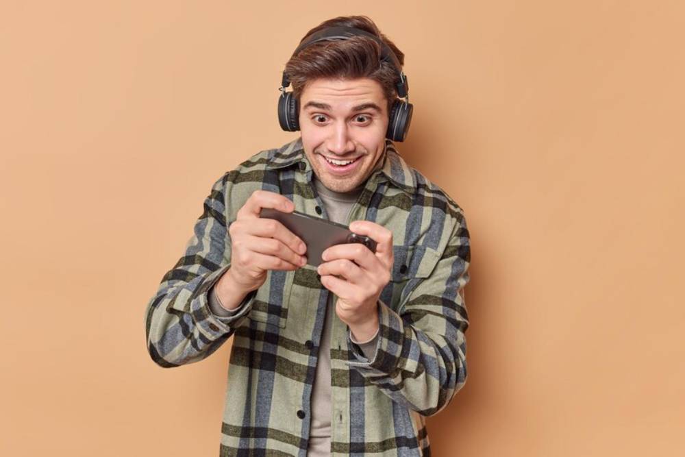 A person wearing headphones is intently playing a mobile game against a plain orange background.