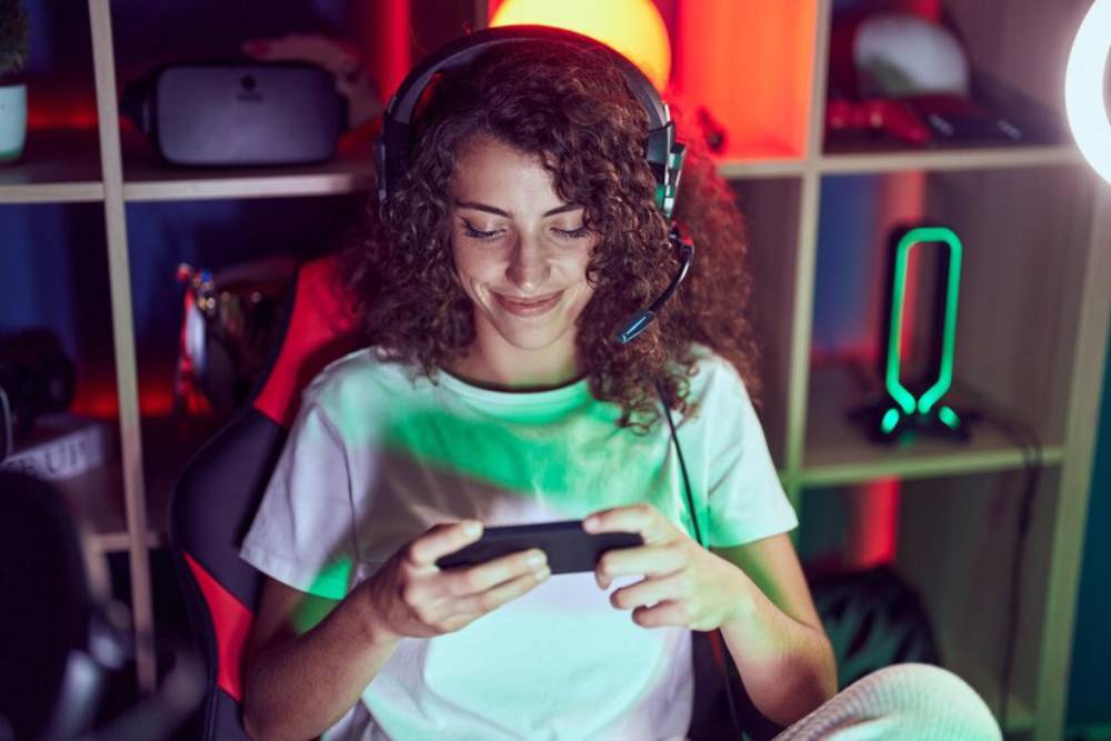 A gamer sits in a colorful, modern room, wearing a headset and focused on their mobile game.